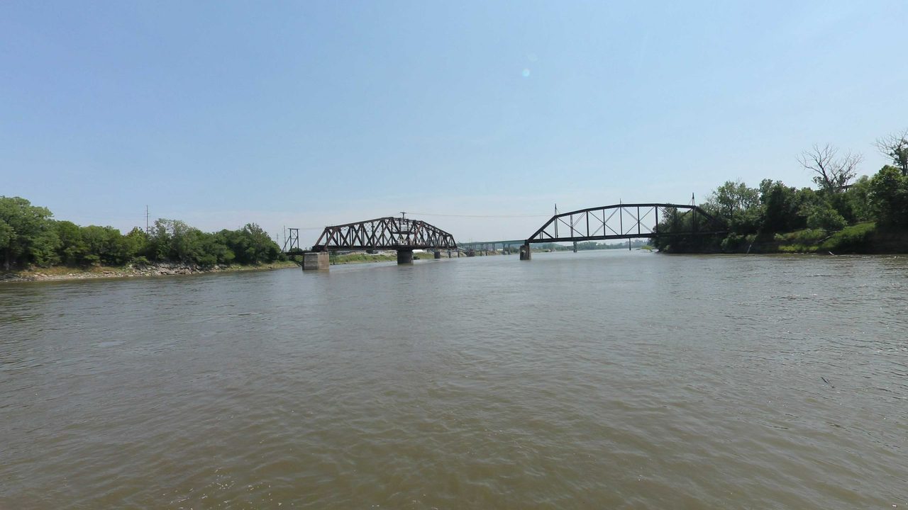 The Missouri River toward two steel truss bridge spans, with wooded banks on both sides, 2023