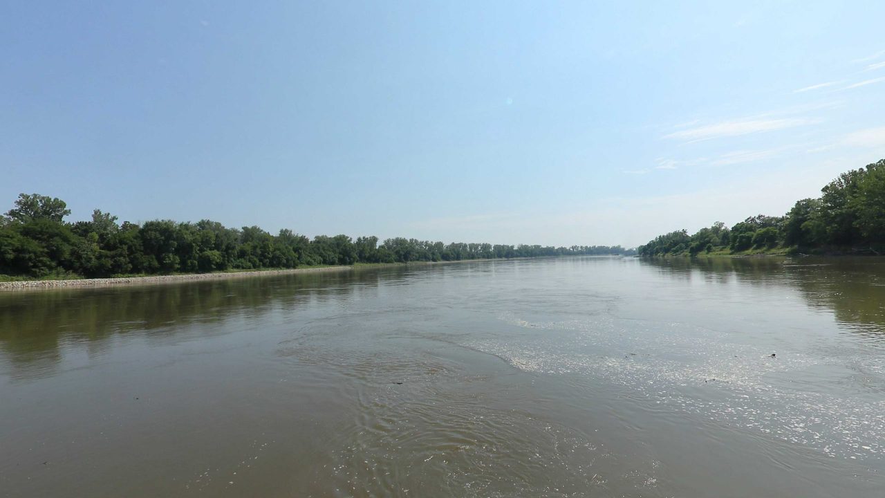 The Missouri River bending past long wooded banks, with bits of driftwood on the water, 2023