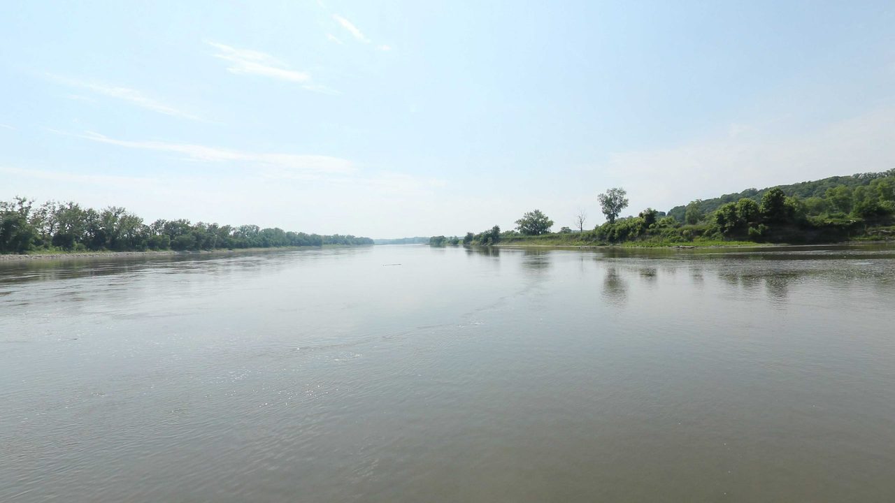 The broad Missouri River between low wooded banks under a pale blue sky, 2023