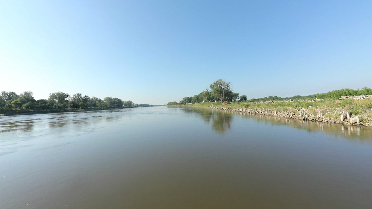 The Missouri River beside a rock-lined bank with a few trees and a house, 2023