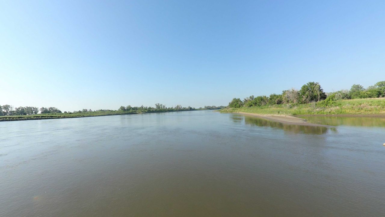 The calm Missouri River with a pale sandbar below a grassy bank, 2023