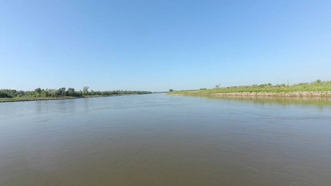 The calm Missouri River between long, low grassy banks, 2023