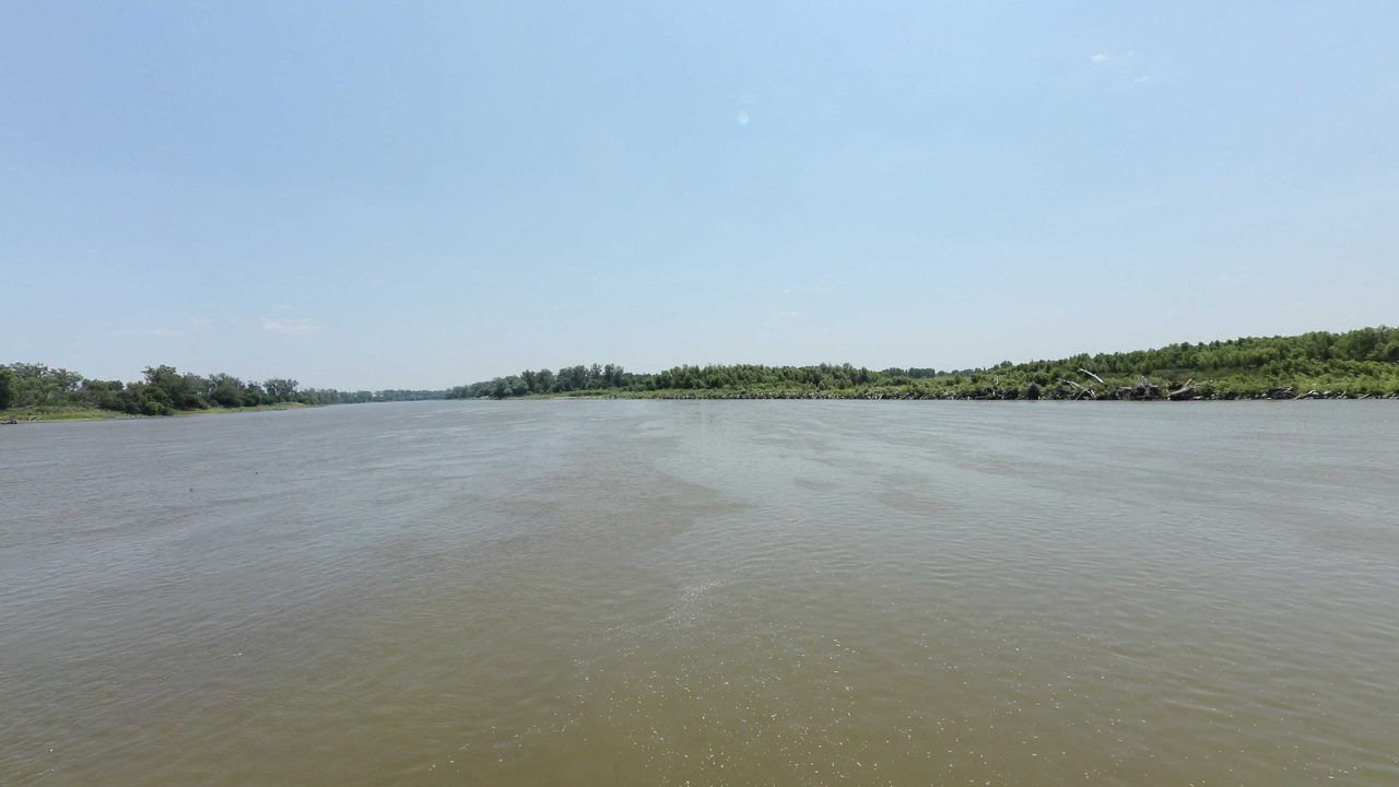 The wide Missouri River under a hazy sky, with a low tree line in the distance, 2023