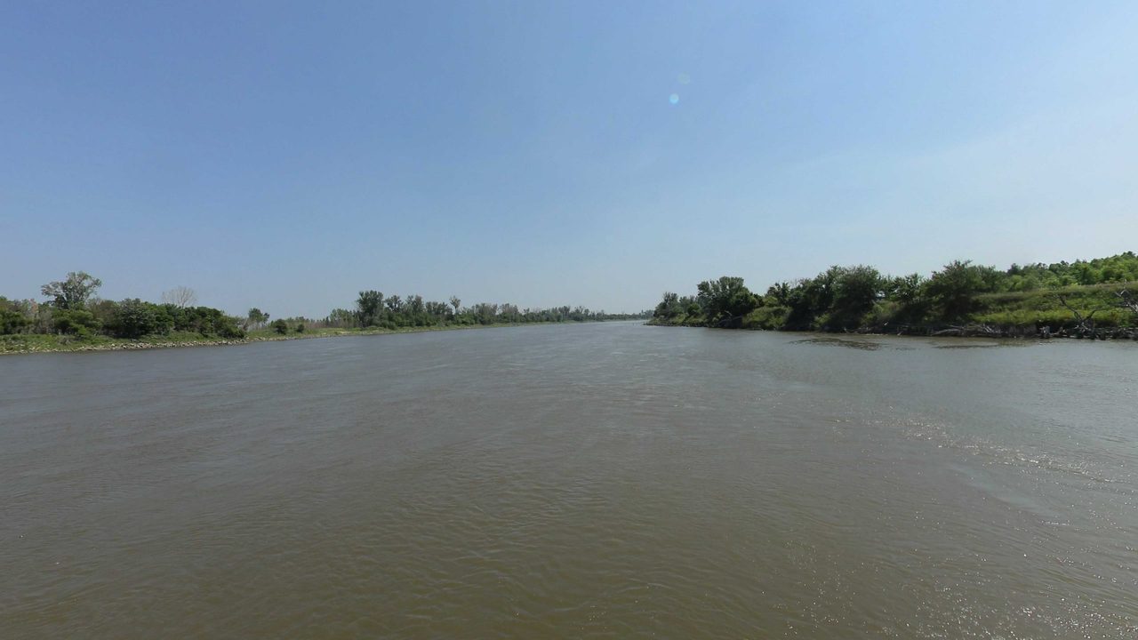 The wide Missouri River between low wooded banks under a clear sky, 2023