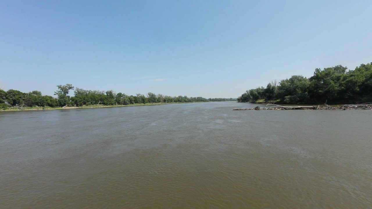 The wide Missouri River between low wooded banks under a clear blue sky, 2023