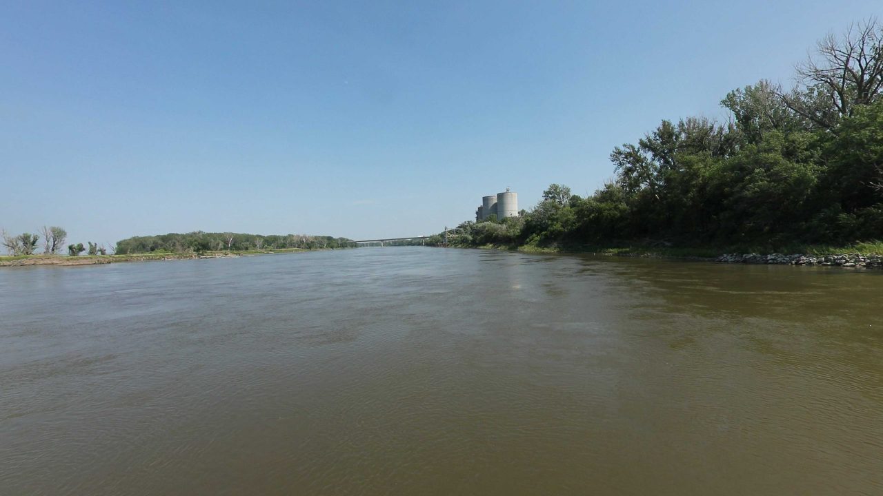 The Missouri River with wooded banks and a distant industrial building on the right, 2023