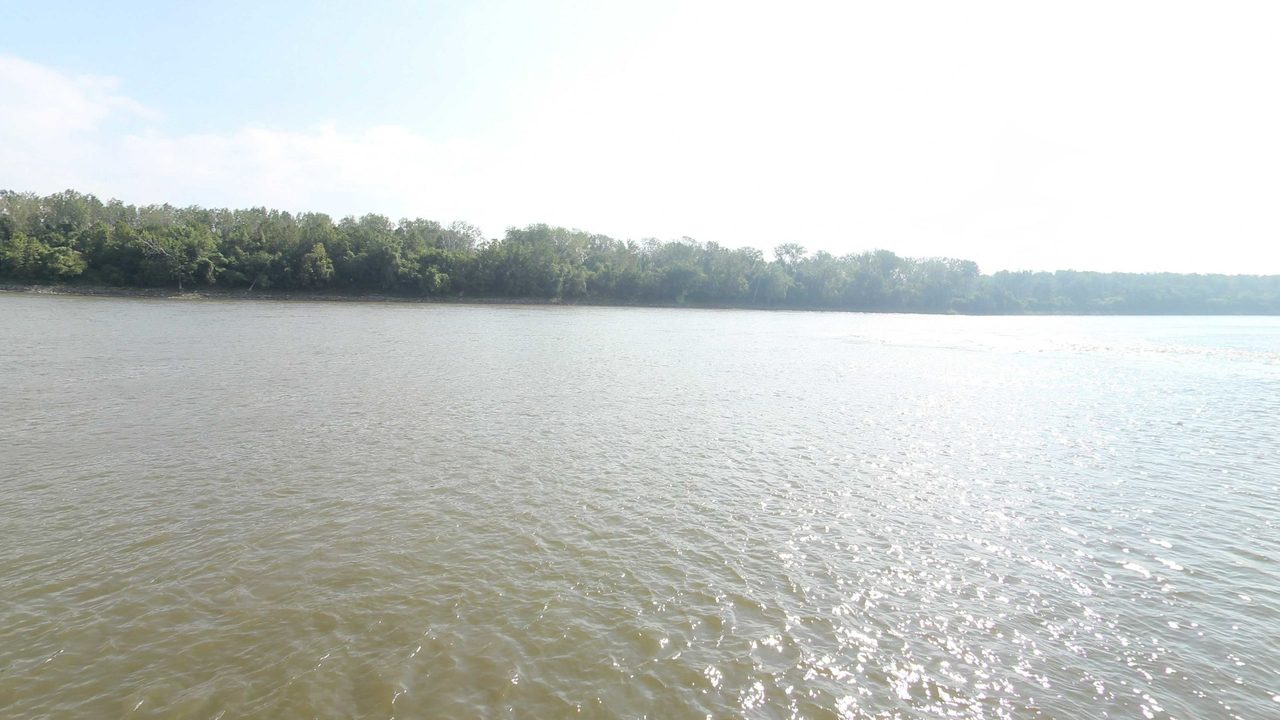 The wide Missouri River with sunlight glittering on the water below a long wooded shore, 2023