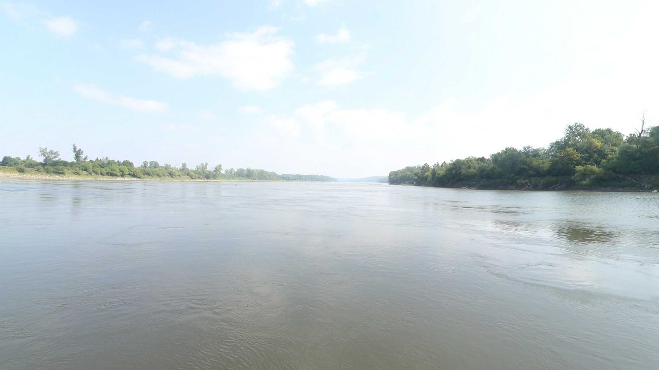 The Missouri River under white clouds, with a low grassy shore on the left and a wooded bank on the right, 2023