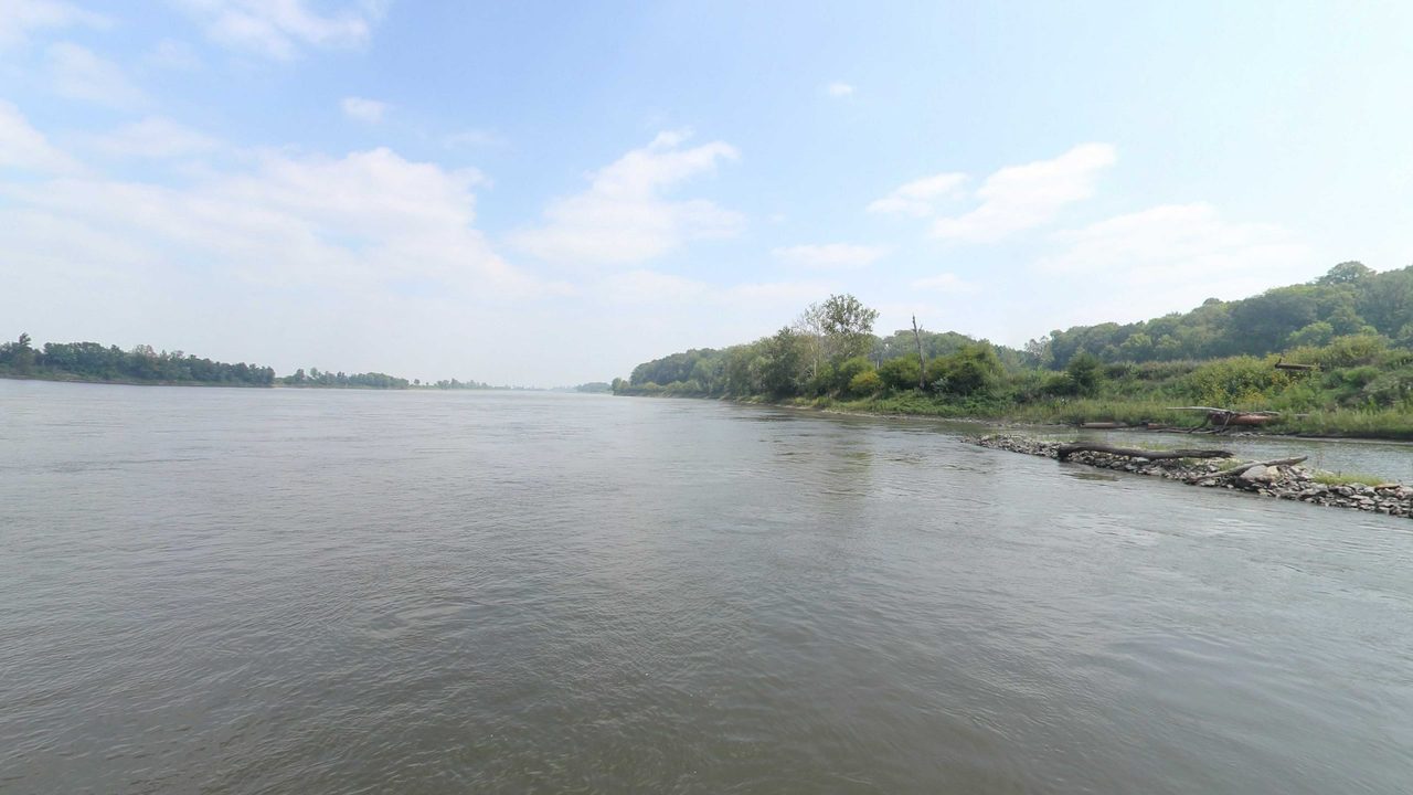 The Missouri River beside a rocky, wooded bank on the right, under scattered white clouds, 2023