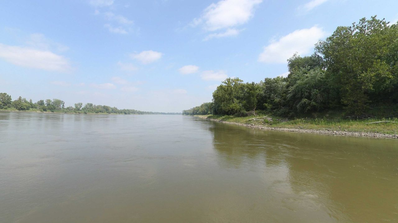 The Missouri River beside a leafy wooded point on the right, under scattered clouds, 2023