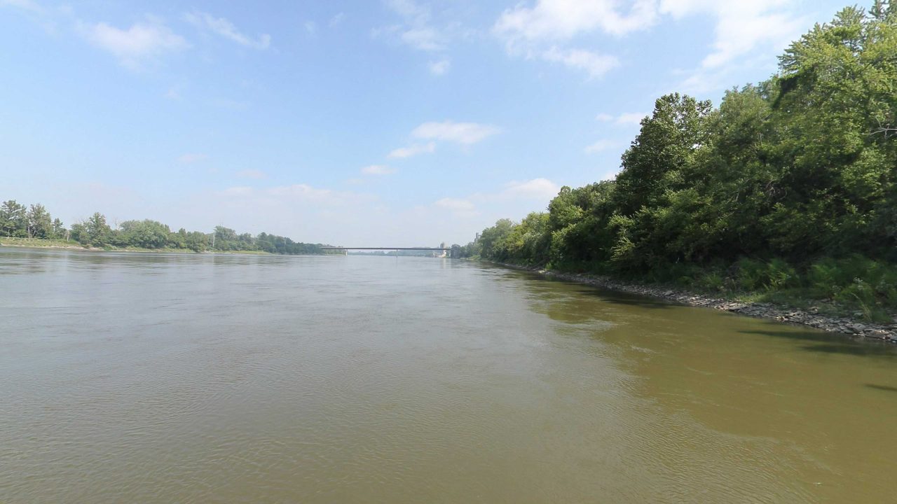 The Missouri River below a leafy wooded bank on the right, under a blue sky with light clouds, 2023