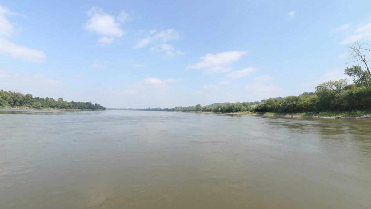 The wide Missouri River between wooded banks under scattered white clouds, 2023