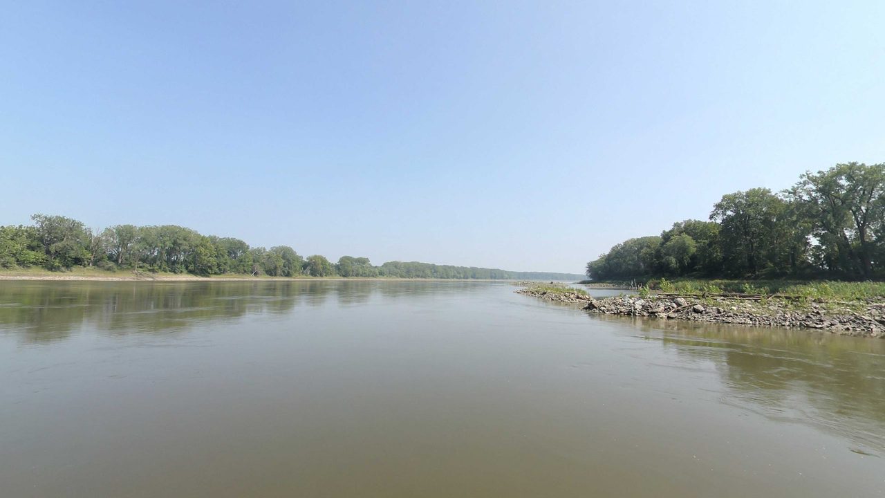 The calm Missouri River beside a rocky point strewn with driftwood, below cottonwoods under a hazy blue sky, 2023