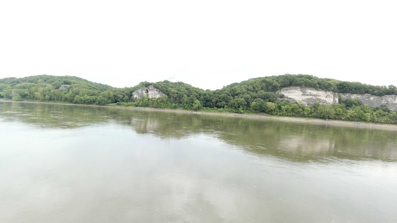 The Missouri River below wooded bluffs with pale limestone cliffs, under a white sky, 2023