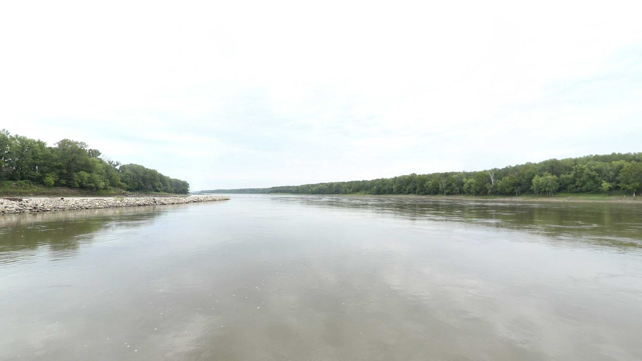 The Missouri River beside a rock-lined bank, with a long wooded shore under a white sky, 2023