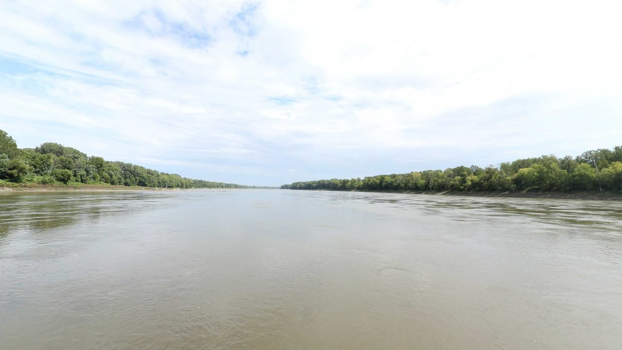 The wide Missouri River between long wooded banks under a sky of white clouds, 2023