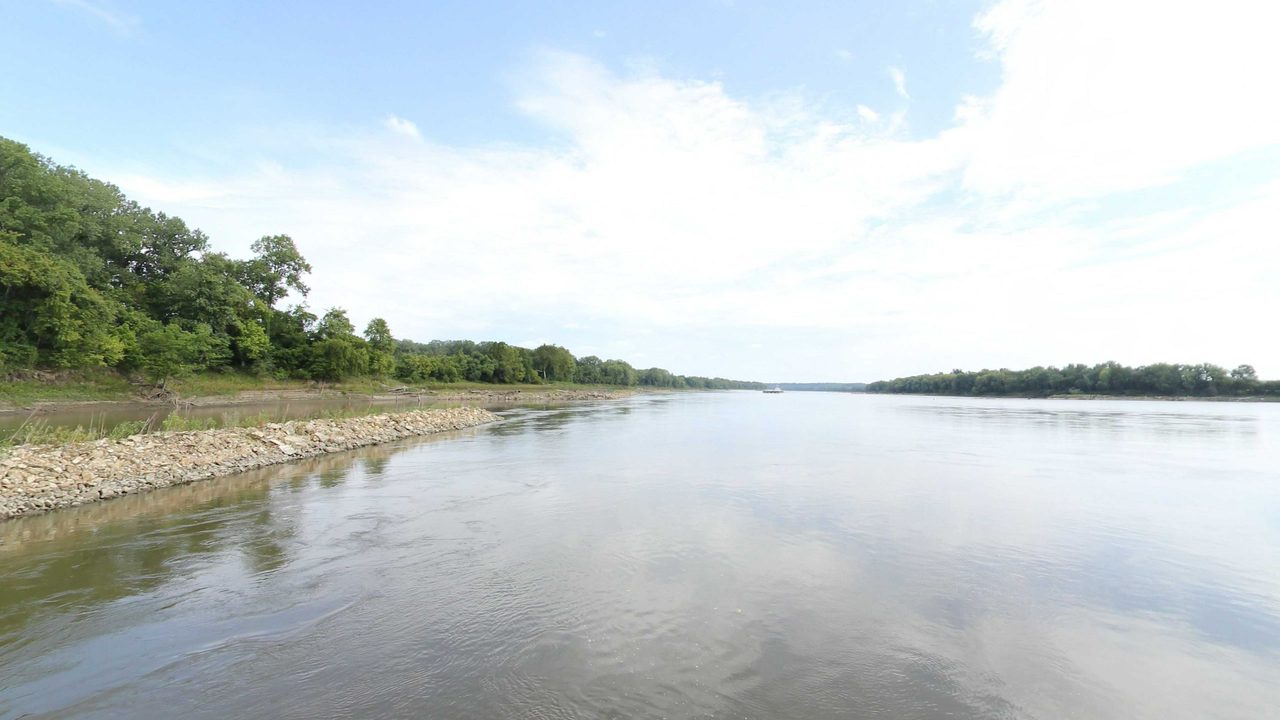The Missouri River beside a rock-lined bank below trees, with a towboat far downstream, 2023