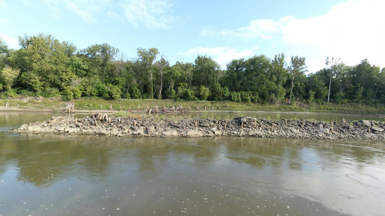 A rock dike with weathered wooden pilings below the wooded bank of the Missouri River, 2023
