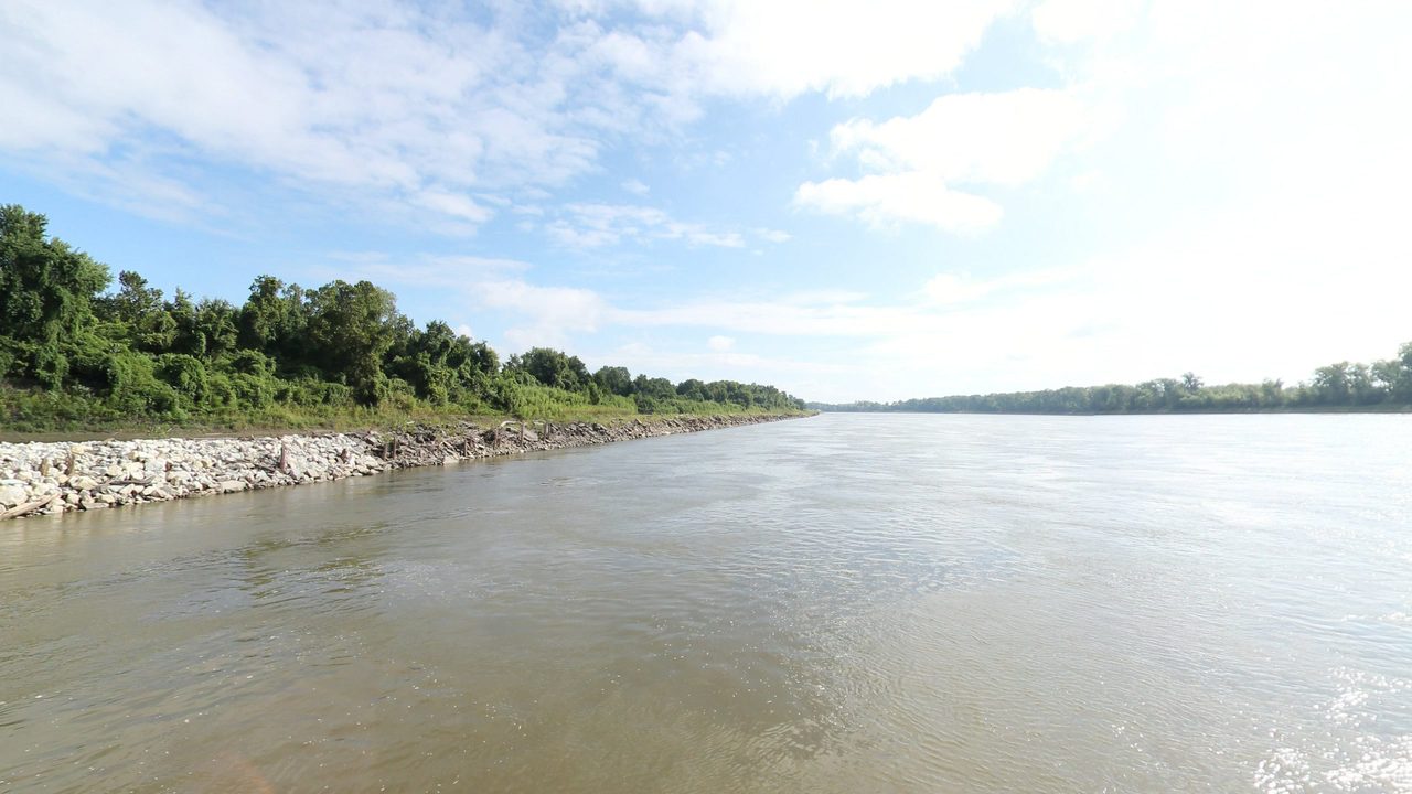 The Missouri River beside a rock-lined bank with trees on the left, under white clouds, 2023