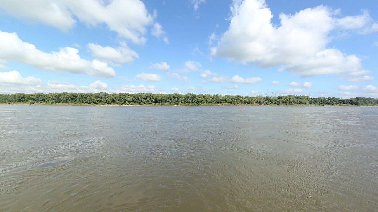 The wide Missouri River below a line of trees and big white clouds, 2023
