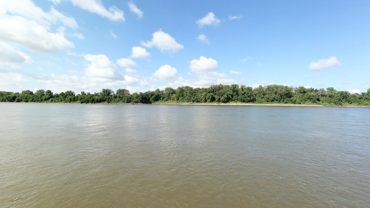 The broad Missouri River below a long wooded shore under scattered white clouds, 2023