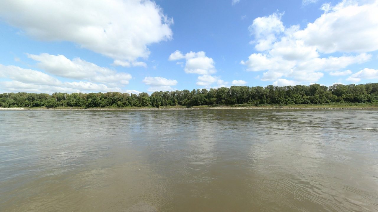 The Missouri River below a long wooded shore with a sandy bar, under billowing clouds, 2023