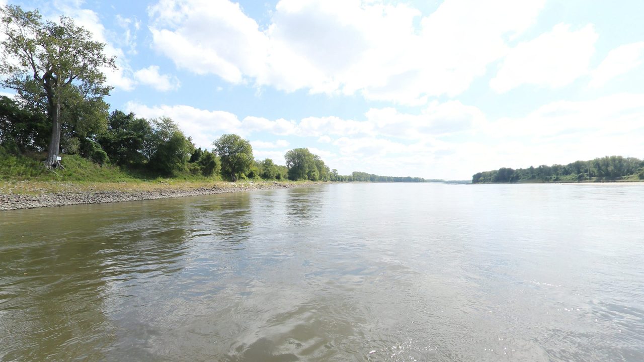 The Missouri River beside a rocky, tree-lined bank under billowing clouds, 2023