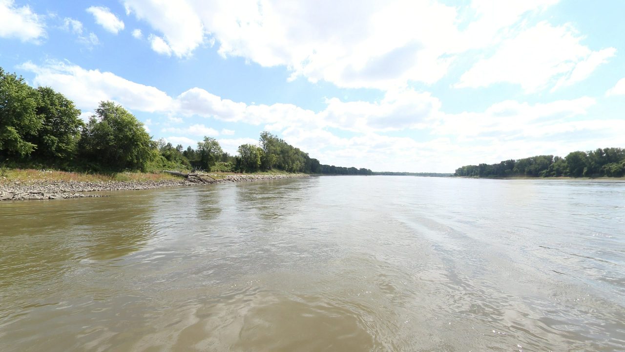The Missouri River beside a rocky, wooded bank on the left, under white clouds, 2023